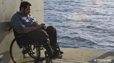 Mann im Rollstuhl steht auf zwei Raedern angelehnt an einer Mauer und schaut in sein Smartphone