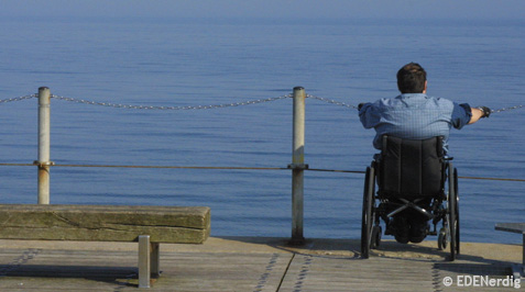 Mann im Rollstuhl steht am Meer, mit dem Ruecken zur Kamera